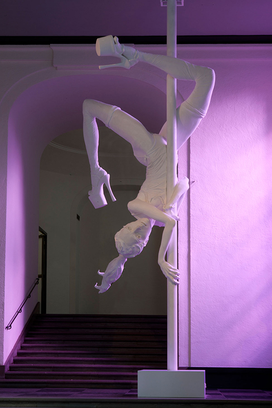 Stor skulptur i vitt av en androgyn person som utför pole dancing och hänger upp och ner på en stolpe.