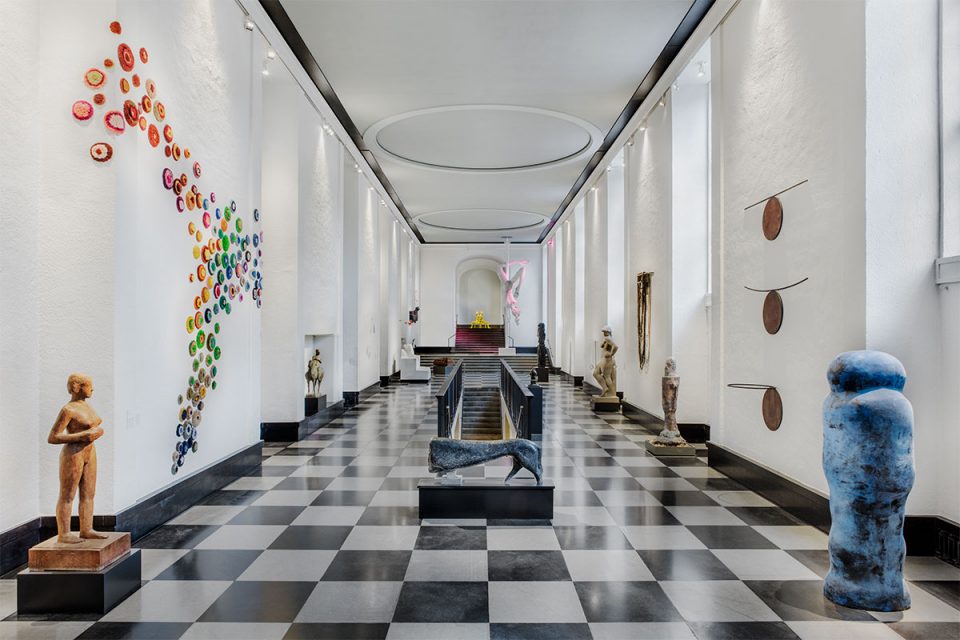 Översikt av Skulpturhallen centrerad vy. Svartvit rutigt golv, trapp till nedre plan i mitten och konstverk placerade på golv och väggar.