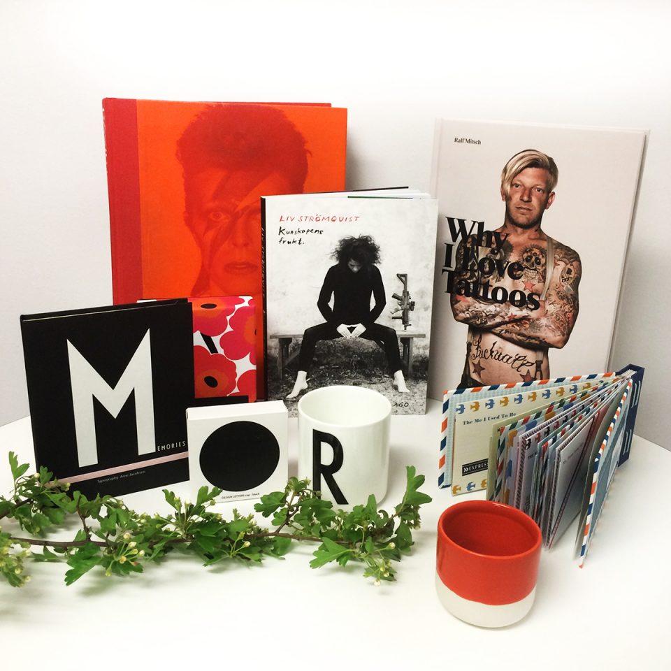 Böcker om David Bowie och tatueringar, vit mugg med svart bokstav på. Röd keramikmugg och en grön kvist.