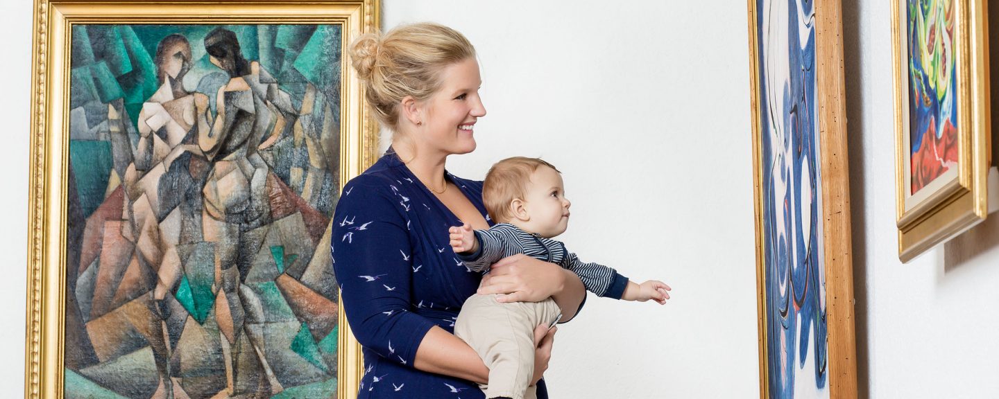Kvinna i blå klänning med barn i famnen som tittar på målningar i museisal.