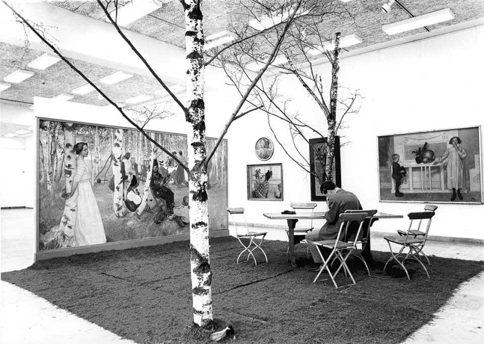 Svartvit foto av museirum med målningar av Carl Larsson i olika format och en björk i förgrunden.